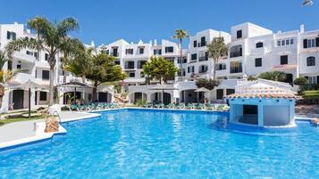 Une piscine entourée de bâtiments blancs dotés de balcons, de palmiers et surplombée d’un ciel bleu azur.
