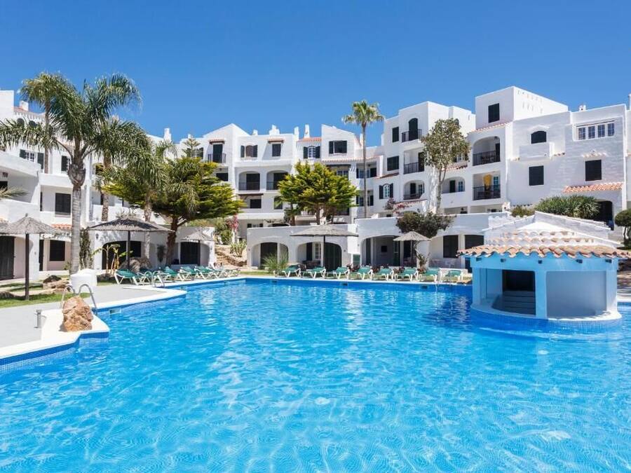 Une piscine entourée de bâtiments blancs dotés de balcons, de palmiers et surplombée d’un ciel bleu azur.