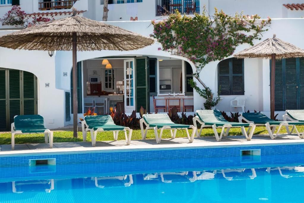 A poolside area with lounge chairs and umbrellas, a pool, and a building with a white facade and green shutters.