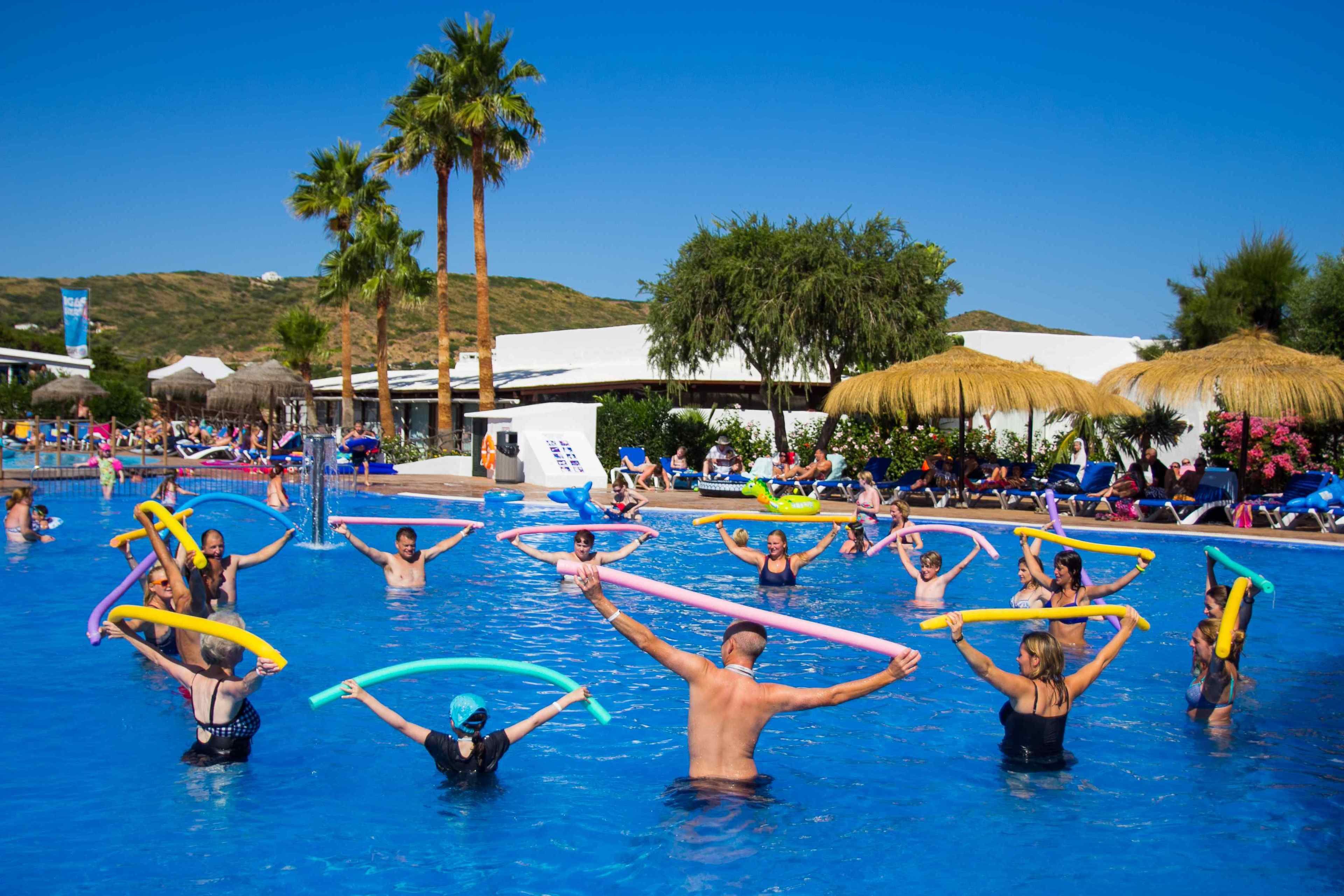 A swimming pool area with people holding colorful noodles, palm trees, and thatched umbrellas in the background.