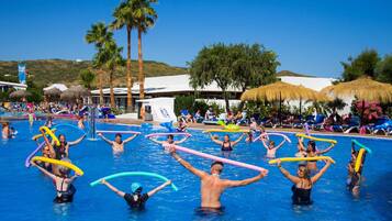 A swimming pool area with people holding colorful noodles, palm trees, and thatched umbrellas in the background.