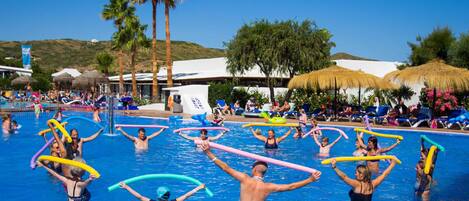A swimming pool area with people holding colorful noodles, palm trees, and thatched umbrellas in the background.
