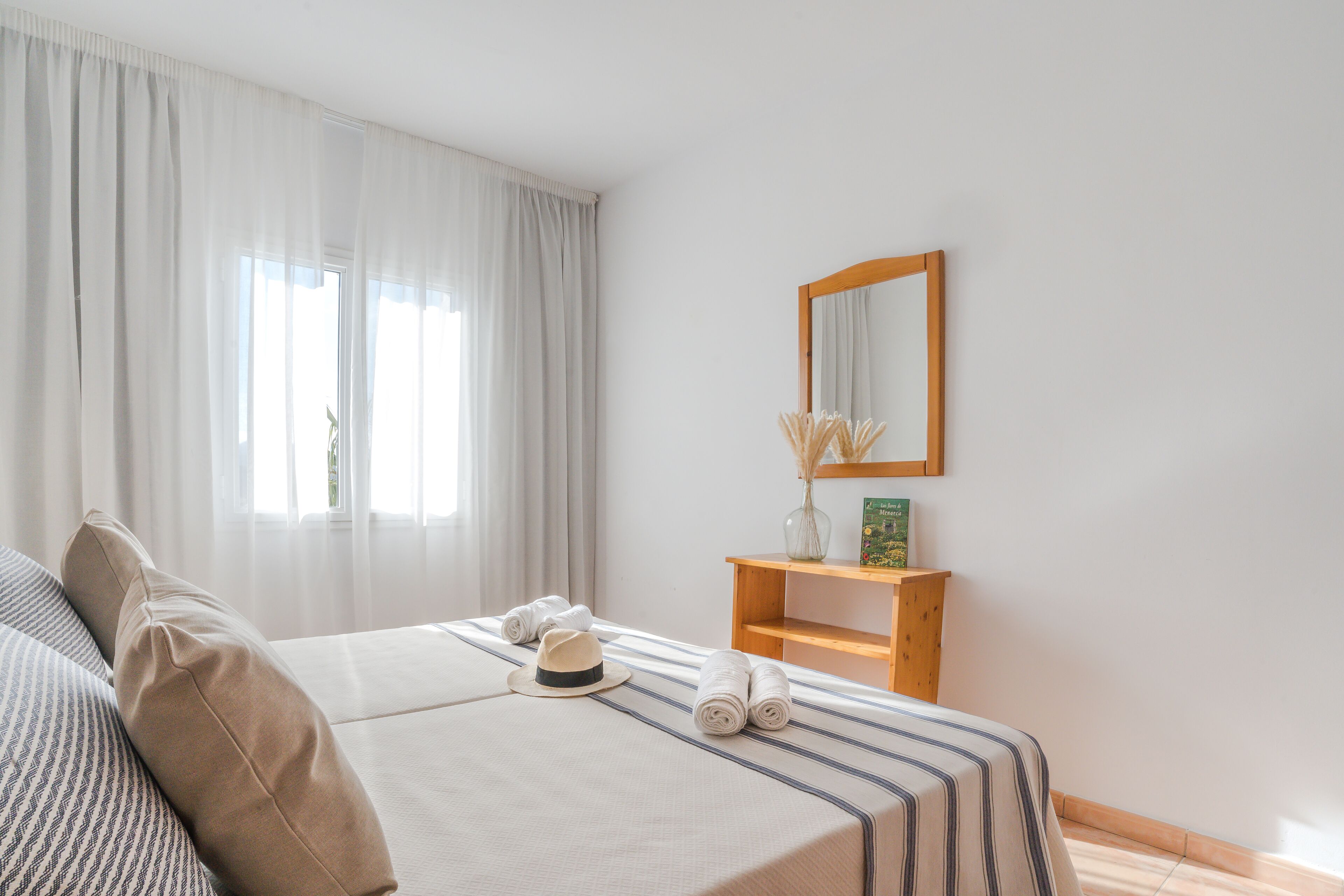 A hotel room with a bed, a wooden nightstand, a mirror, and a window with curtains.