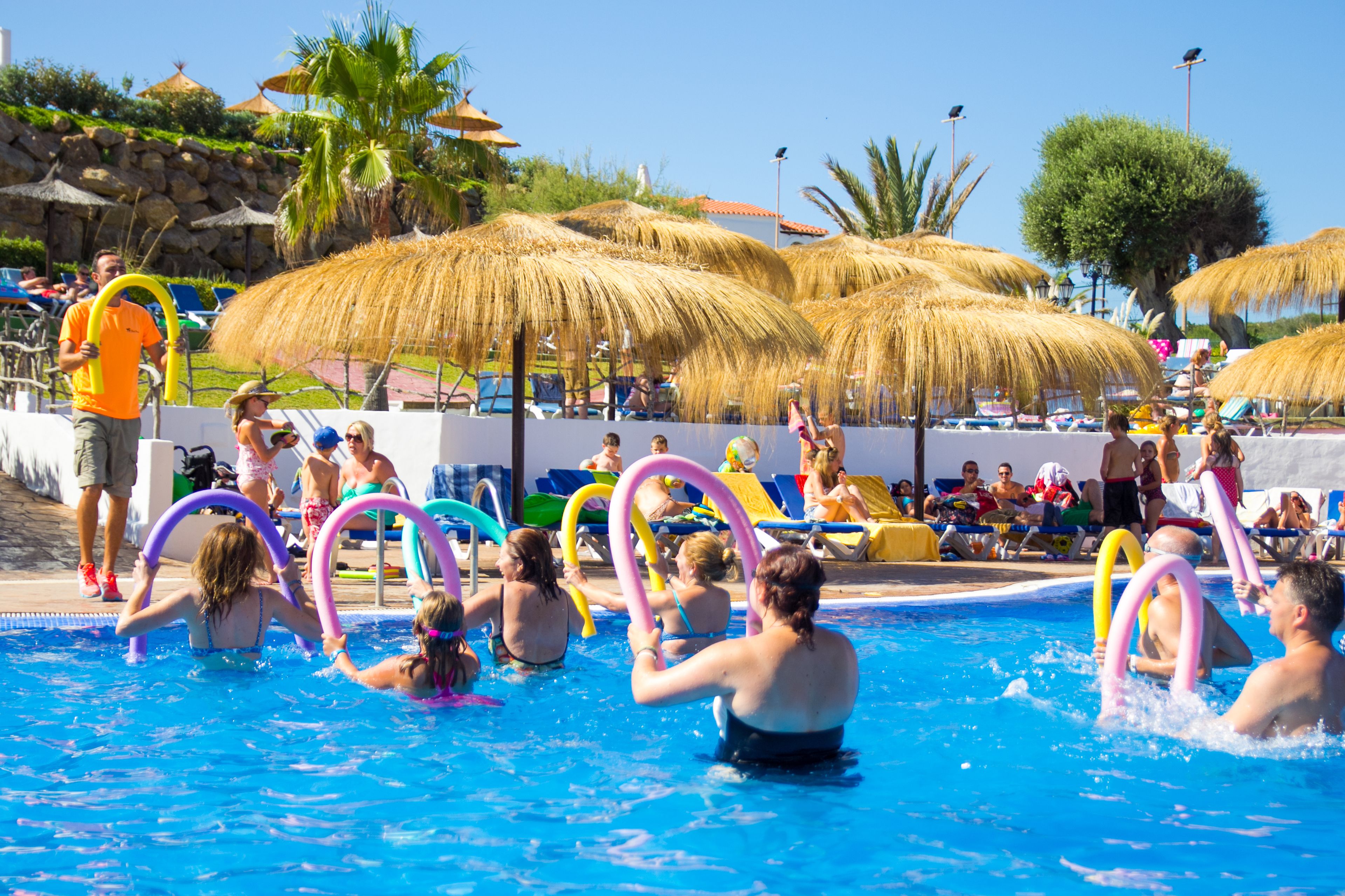 A swimming pool area with multiple people, some holding colorful rings, and thatched umbrellas providing shade.