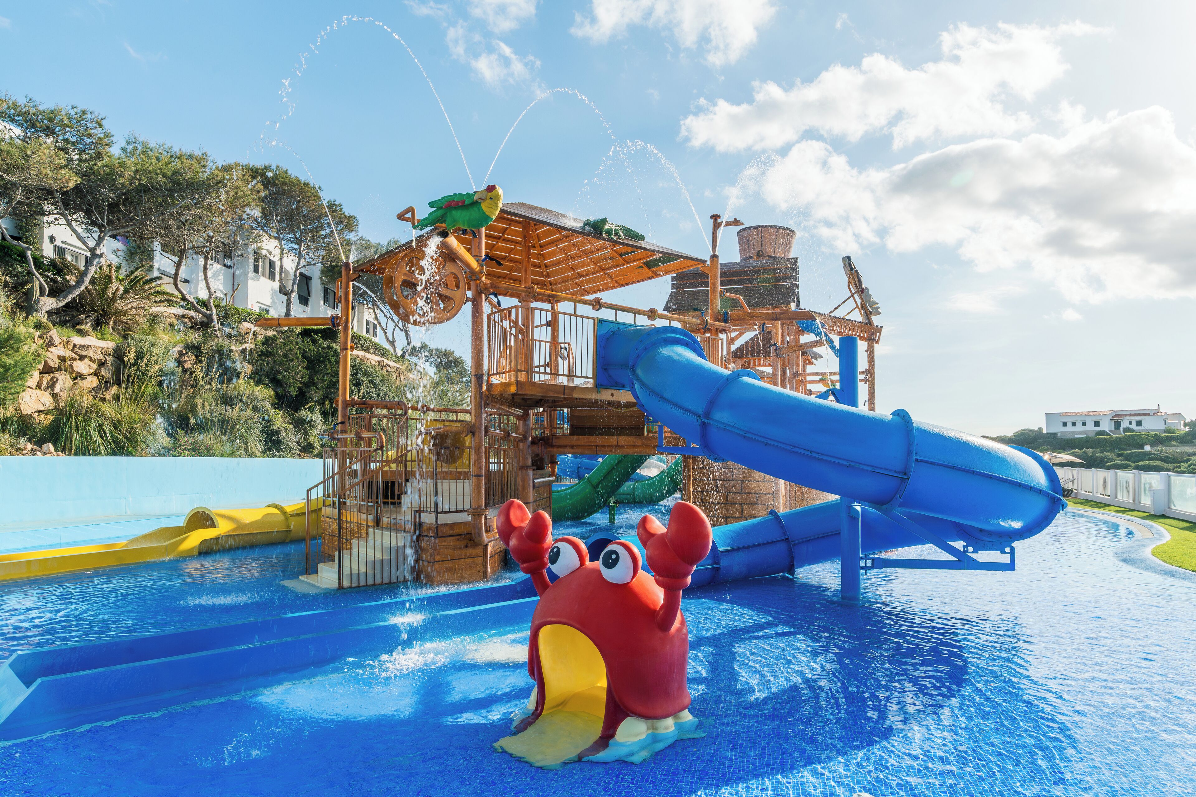 Un parque acuático con un tobogán azul, una estructura de juego de madera y una gran decoración de cangrejo en la piscina.