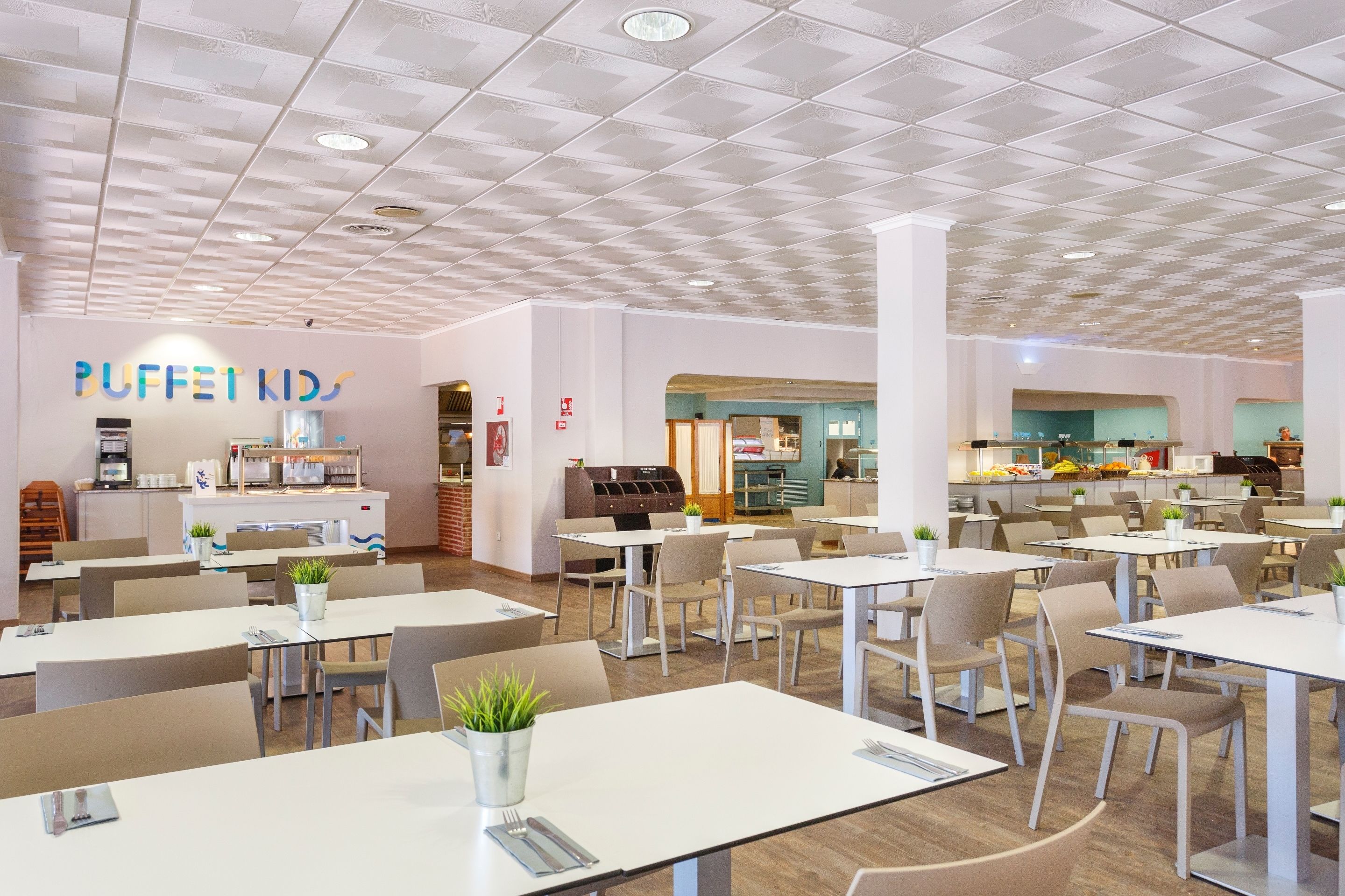 A modern dining area with white tables and chairs, a buffet counter, and a kitchen in the background.