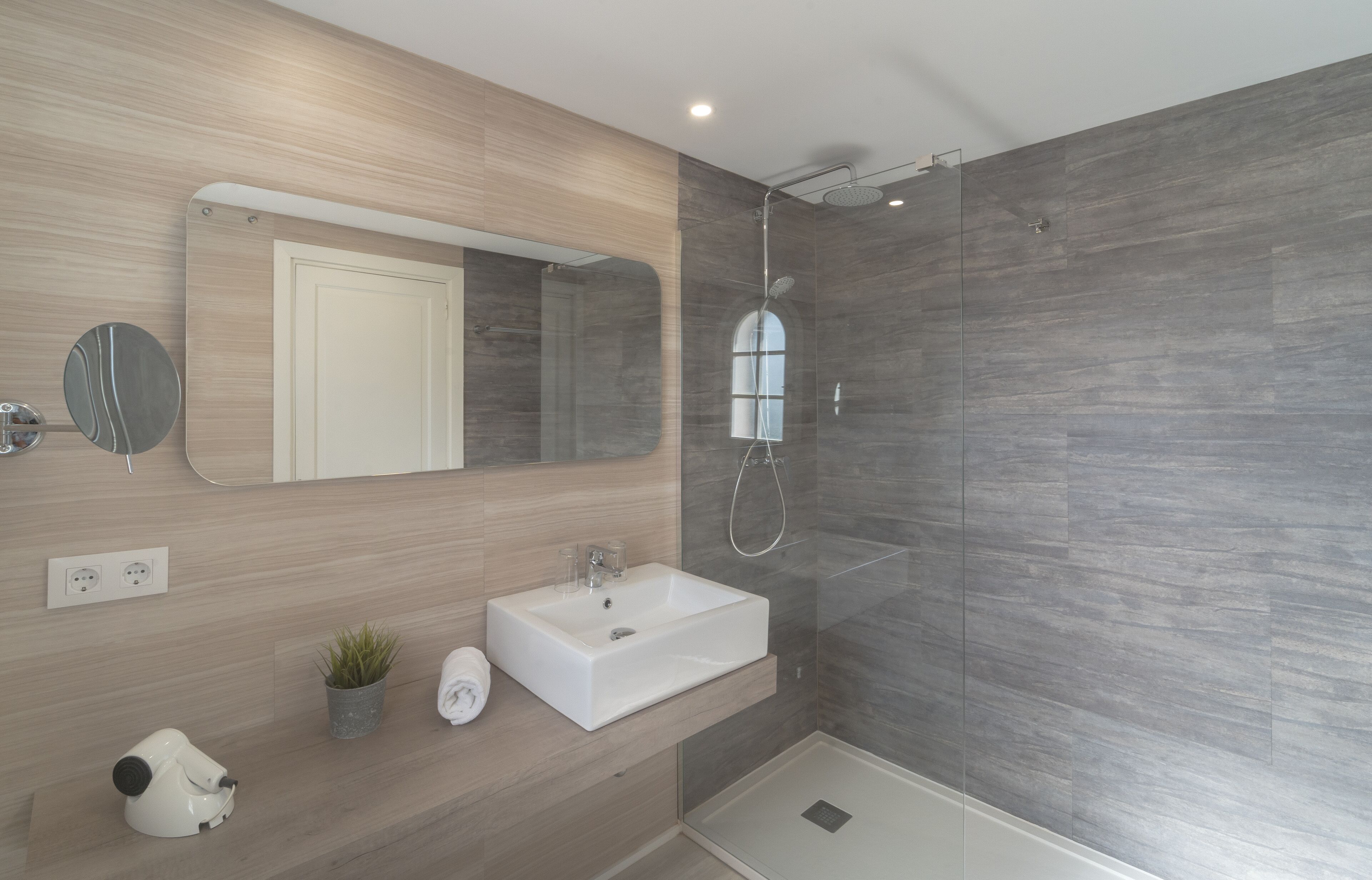 A modern bathroom with a large mirror, a sink, a shower, and a potted plant.