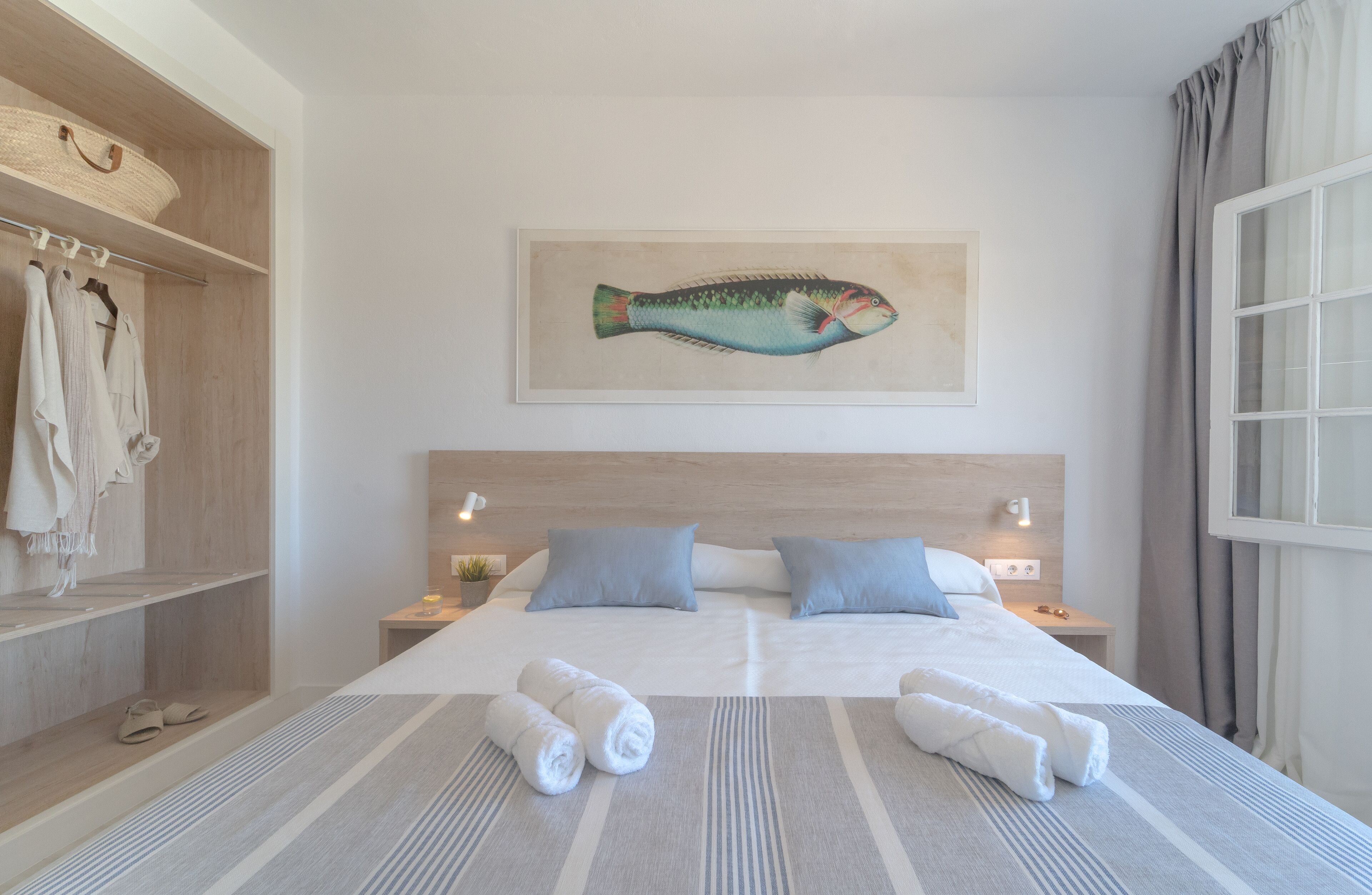 A hotel room with a bed, two pillows, a nightstand, a lamp, a wall-mounted fish painting, a window with curtains, and a wardrobe with hanging clothes.
