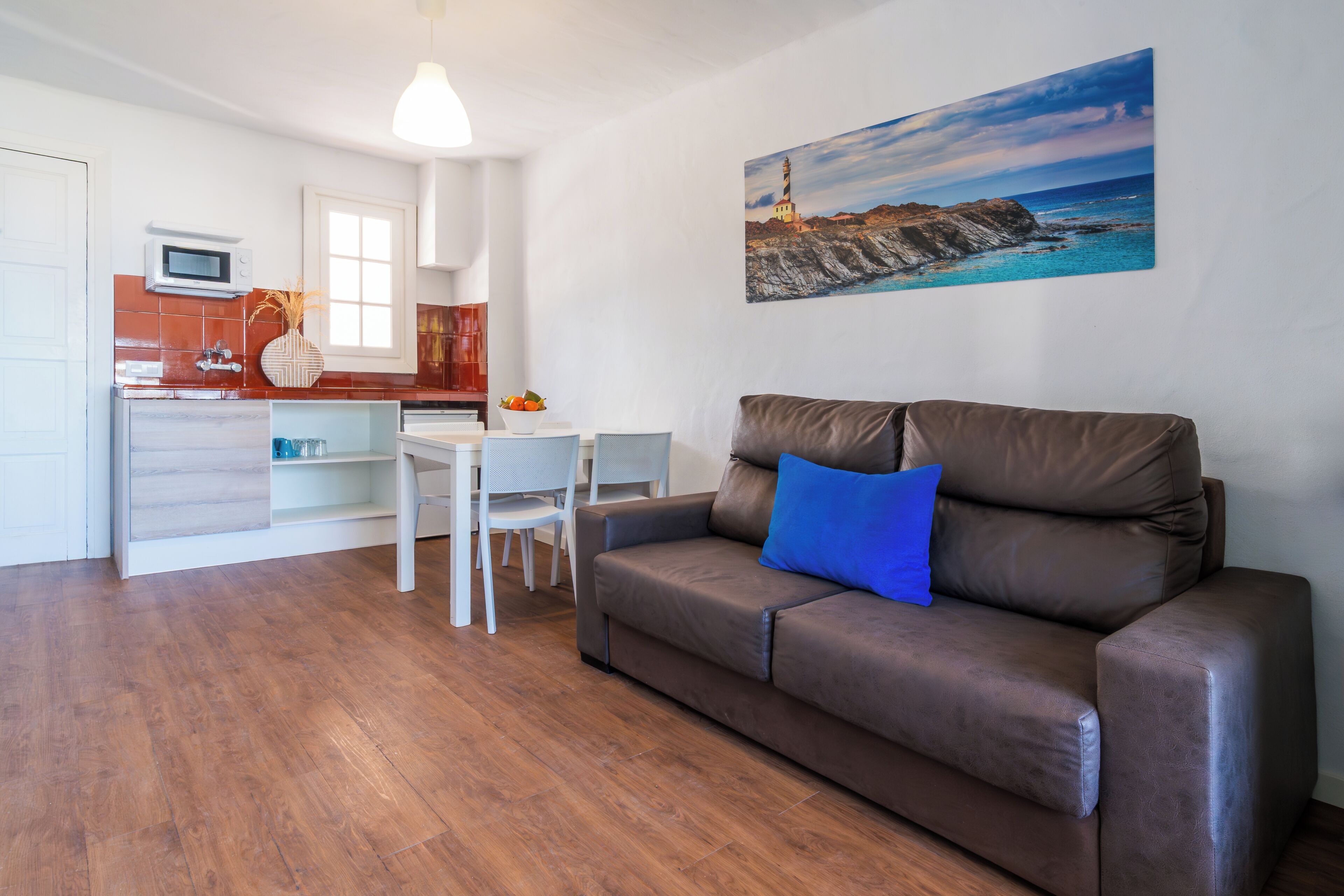 A modern living room with a brown leather sofa, a white dining table, and a kitchen area with a microwave.