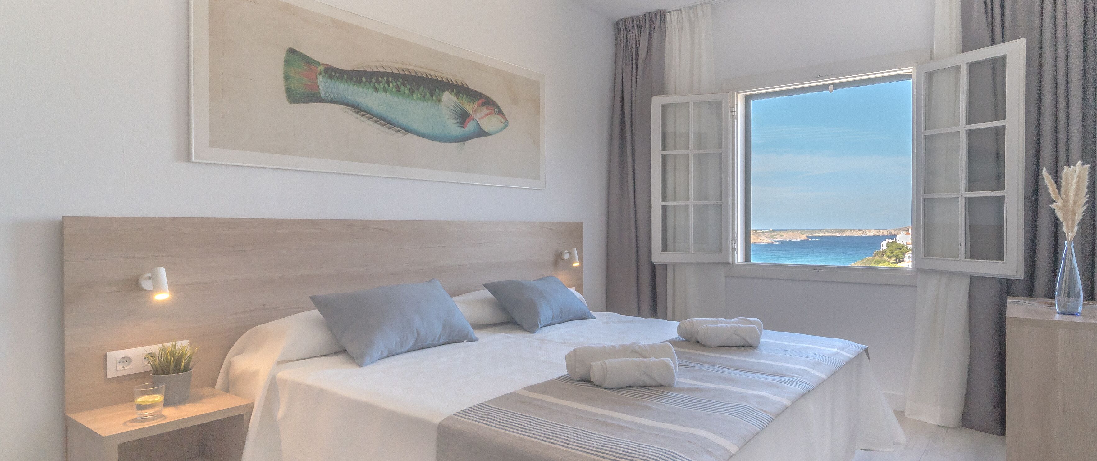 Quarto de hotel com uma cama grande, cabeceira, pintura de um peixe, janela com cortinas e vista para o mar.