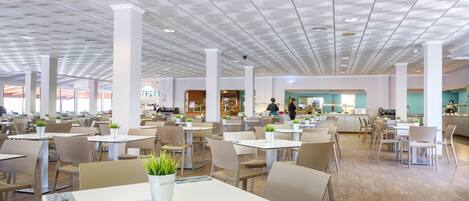 A spacious cafeteria with multiple tables and chairs, white pillars, and ceiling lights.