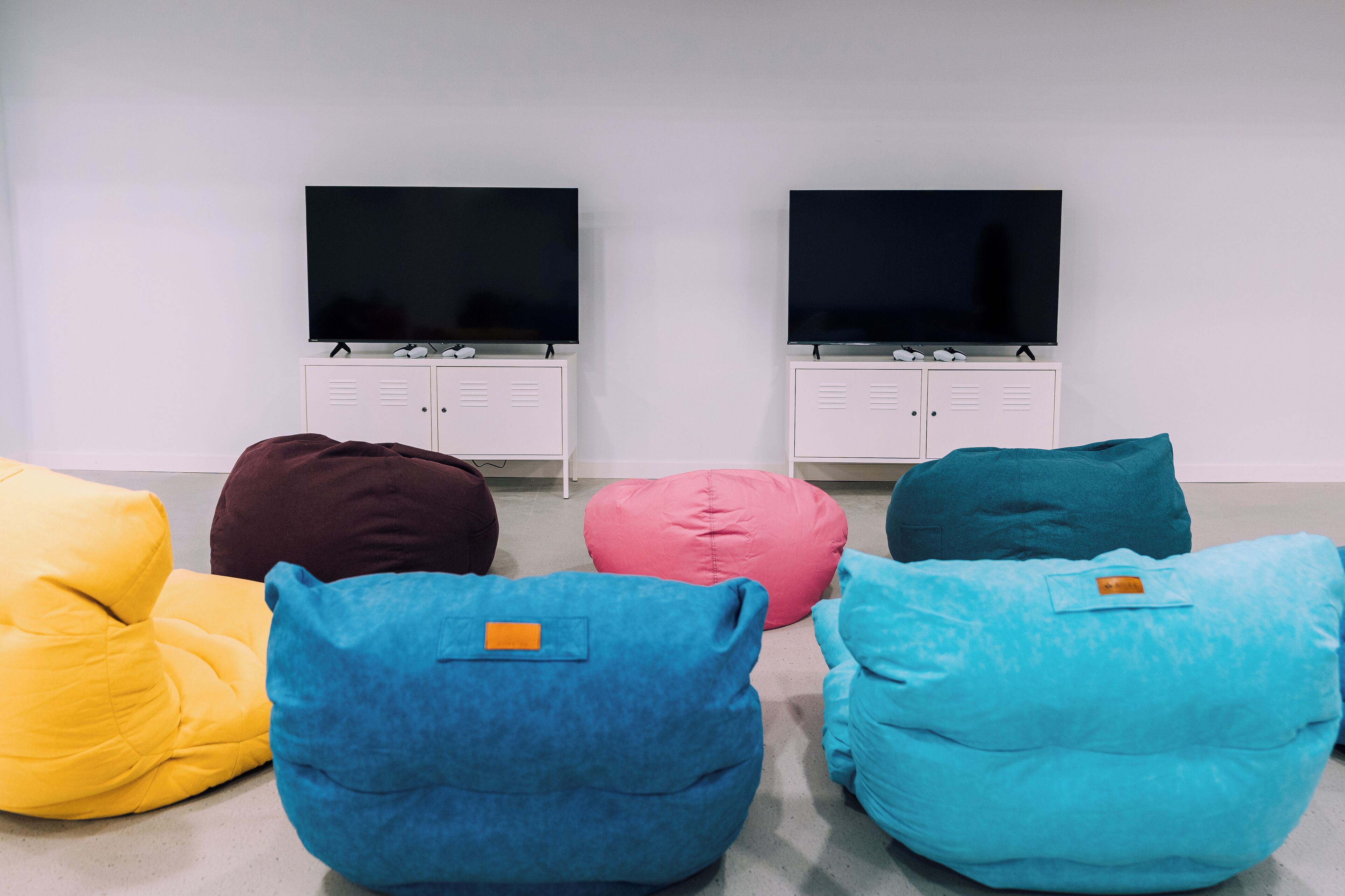 Una habitación con dos televisores sobre muebles blancos, una pared blanca y cinco pufs en amarillo, azul, rosa, verde azulado y azul oscuro.