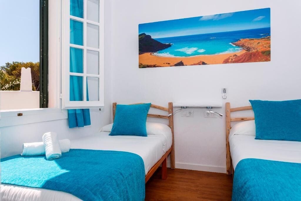 Una habitación con dos camas, cada una con cubrecama y almohada azules, un cuadro de una playa en la pared y una ventana con cortinas azules.