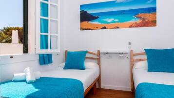 Una habitación con dos camas, cada una con cubrecama y almohada azules, un cuadro de una playa en la pared y una ventana con cortinas azules.