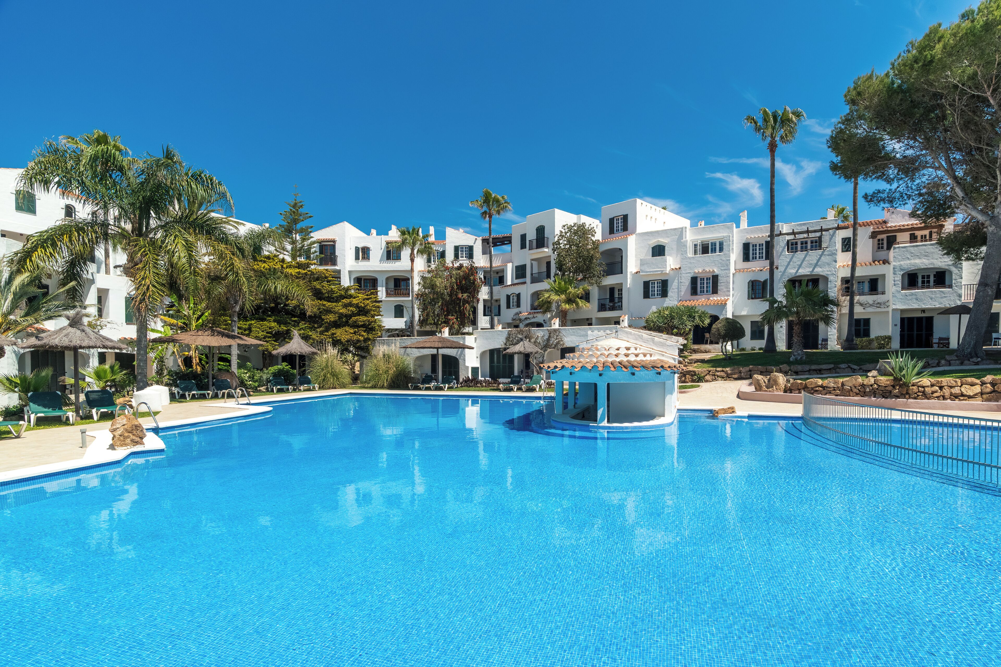 Una gran piscina con un mirador, rodeada de edificios blancos con balcones y palmeras.
