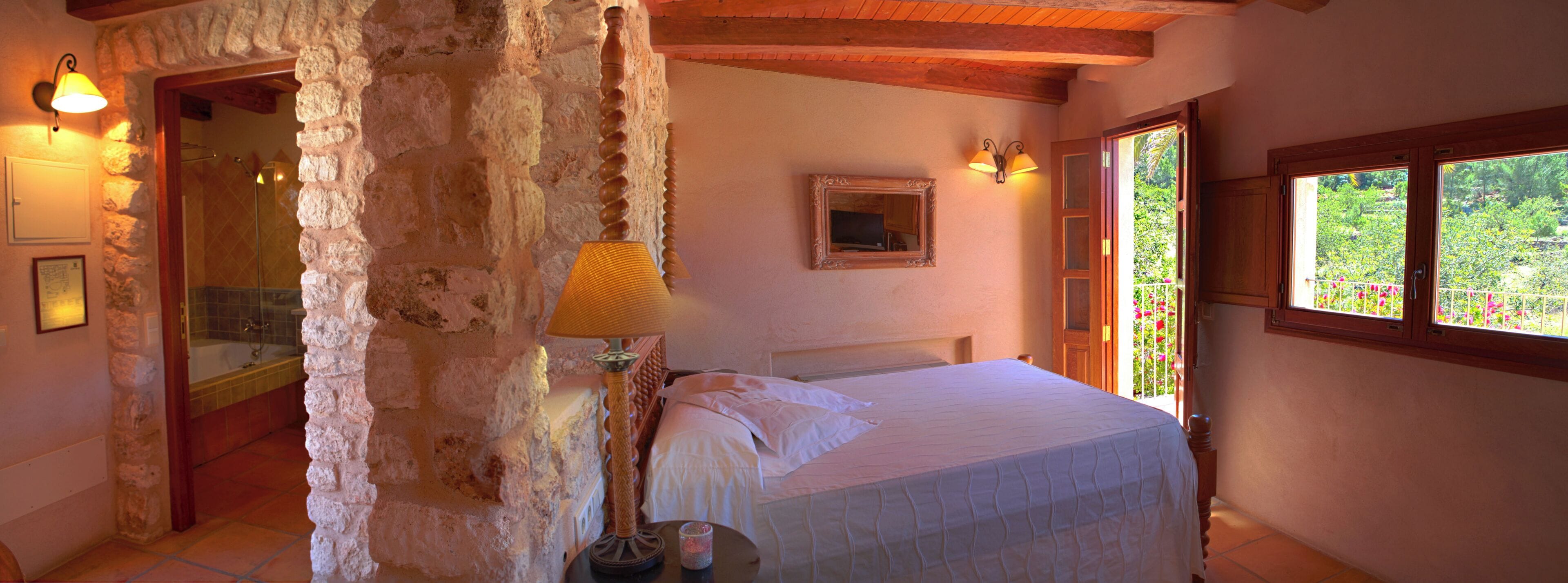 A bedroom with a stone wall, a bed, a window with a view of greenery, and a bathroom with a shower and a tub.