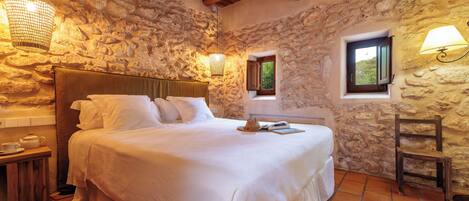 A bedroom with a stone wall, wooden ceiling beams, a bed with white linens, a bedside table with a teapot, and a small window with a view of greenery.