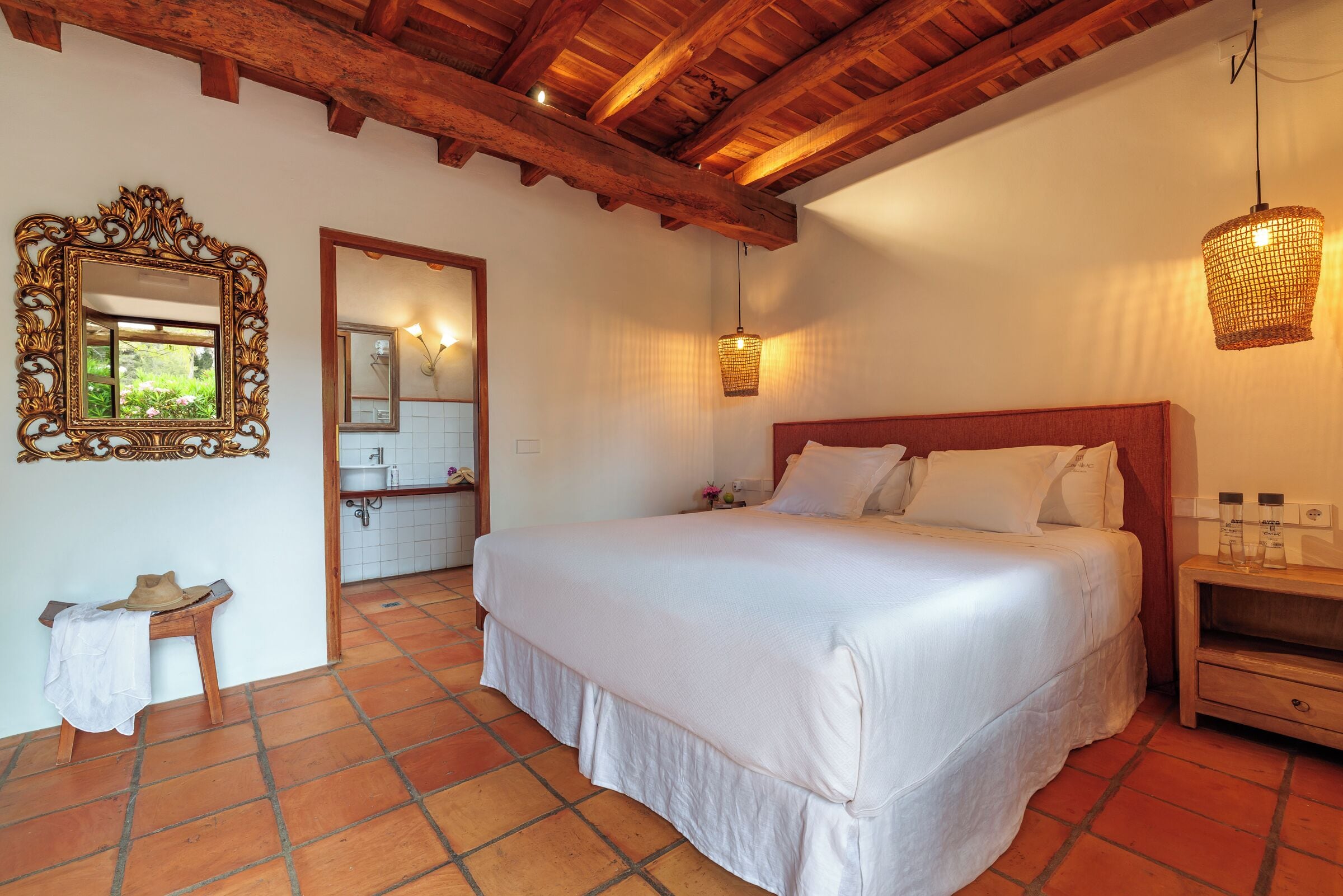 A bedroom with a large bed, wooden ceiling beams, a mirror, and a hanging light fixture.