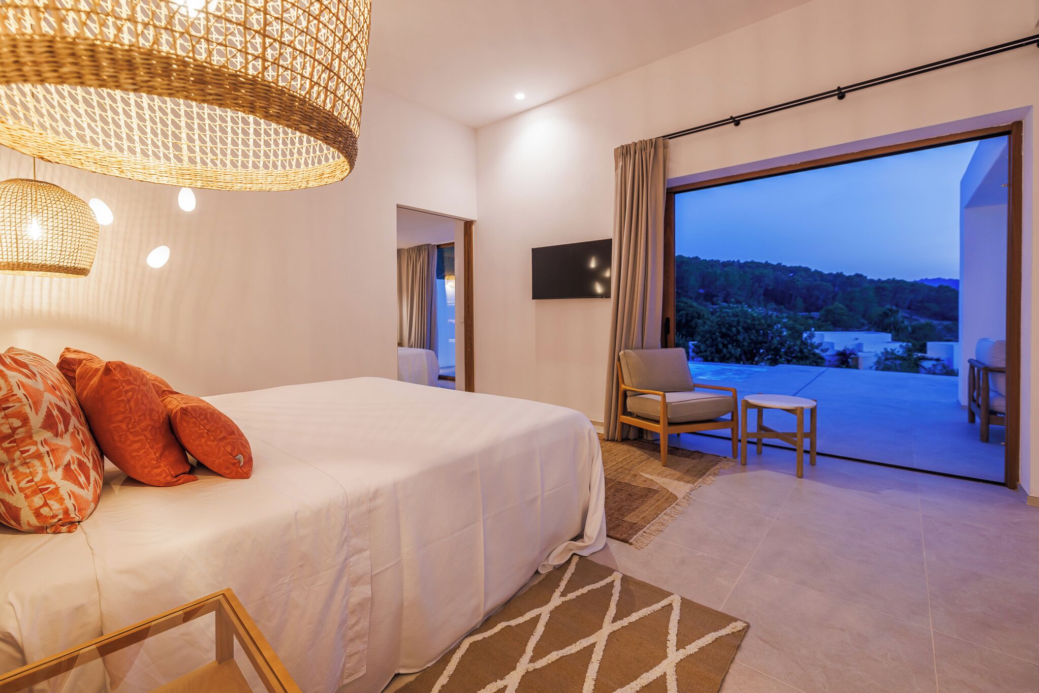 A bedroom with a large bed, a chair, a small table, a TV, and a balcony with a view.