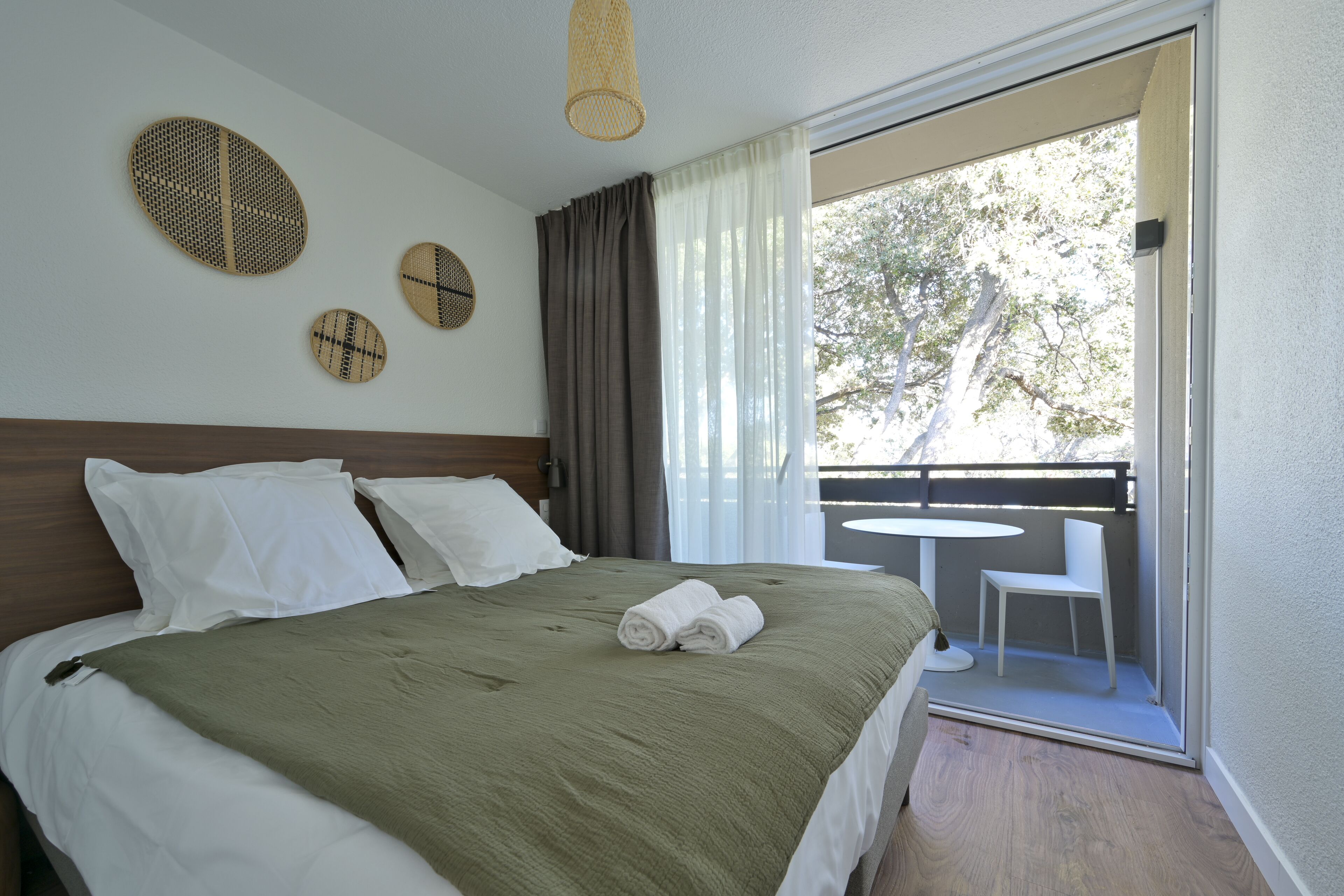 A neatly made bed with a green blanket, white pillows, and two rolled towels. There are three woven wall decorations and a balcony with a white chair and table.