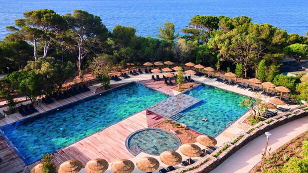 A large outdoor swimming pool with multiple lanes, surrounded by lounge chairs and umbrellas, set against a backdrop of trees and the sea. - Belambra Clubs Presqu'île de Giens - Les Criques (Hyères)