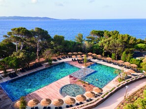 Une grande piscine extérieure avec plusieurs couloirs de nage, entourée de chaises longues et de parasols, le tout dans un cadre verdoyant avec des arbres et la mer en toile de fond.