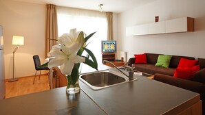 A modern kitchen with a sink, a dining area with a chair, a table, and a vase with flowers. There is a brown sofa with red and green cushions in the living area. - Akacfa Holiday Apartments (Budapest)