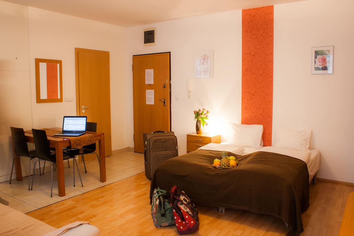 A hotel room with a bed, a desk with a laptop, a suitcase, and a vase with flowers.