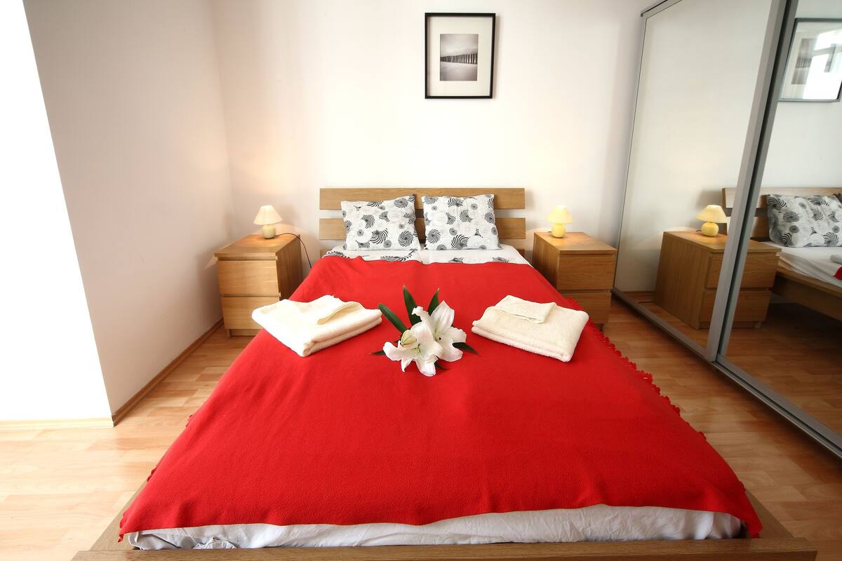 A neatly made bed with a red blanket, two bedside tables with lamps, a mirror, and a framed picture on the wall.