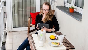 A person sitting at a wooden table on a balcony with a white tablecloth, a teapot, a cup, a plate with food, and a potted plant. - Akacfa Holiday Apartments (Budapest)