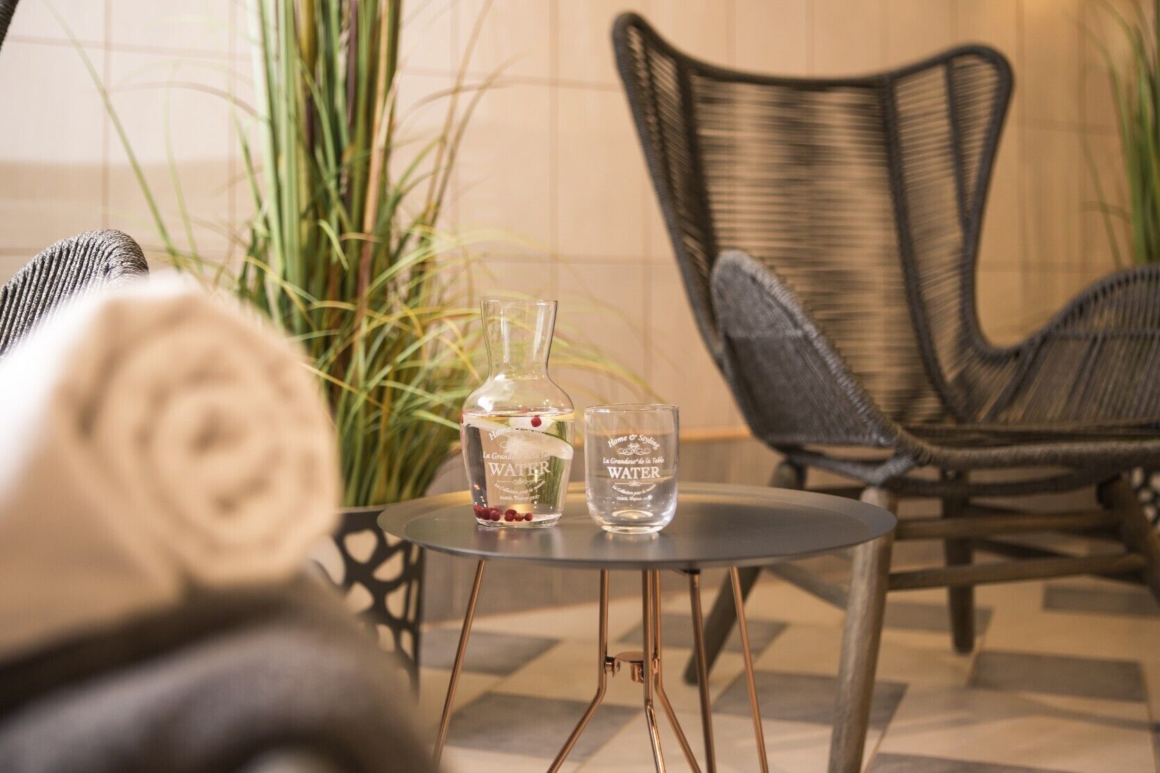 A round table with a water bottle and a glass, a wicker chair, and a rolled towel.