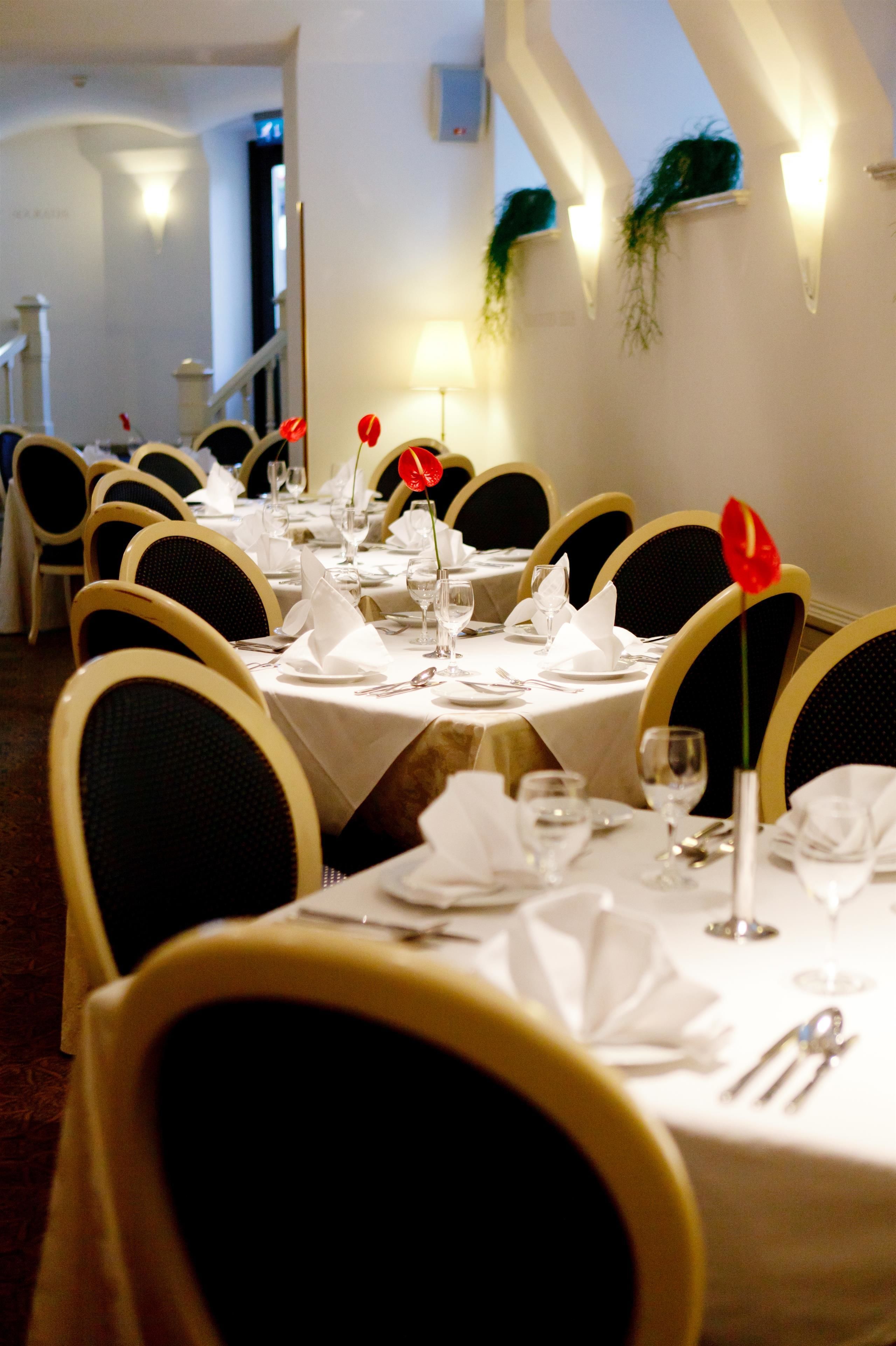 Comedor con mesas redondas dispuestas para cuatro comensales, cada una con mantel blanco, cubiertos y centros de mesa con flores rojas.