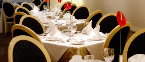 Comedor con mesas redondas dispuestas para cuatro comensales, cada una con mantel blanco, cubiertos y centros de mesa con flores rojas.