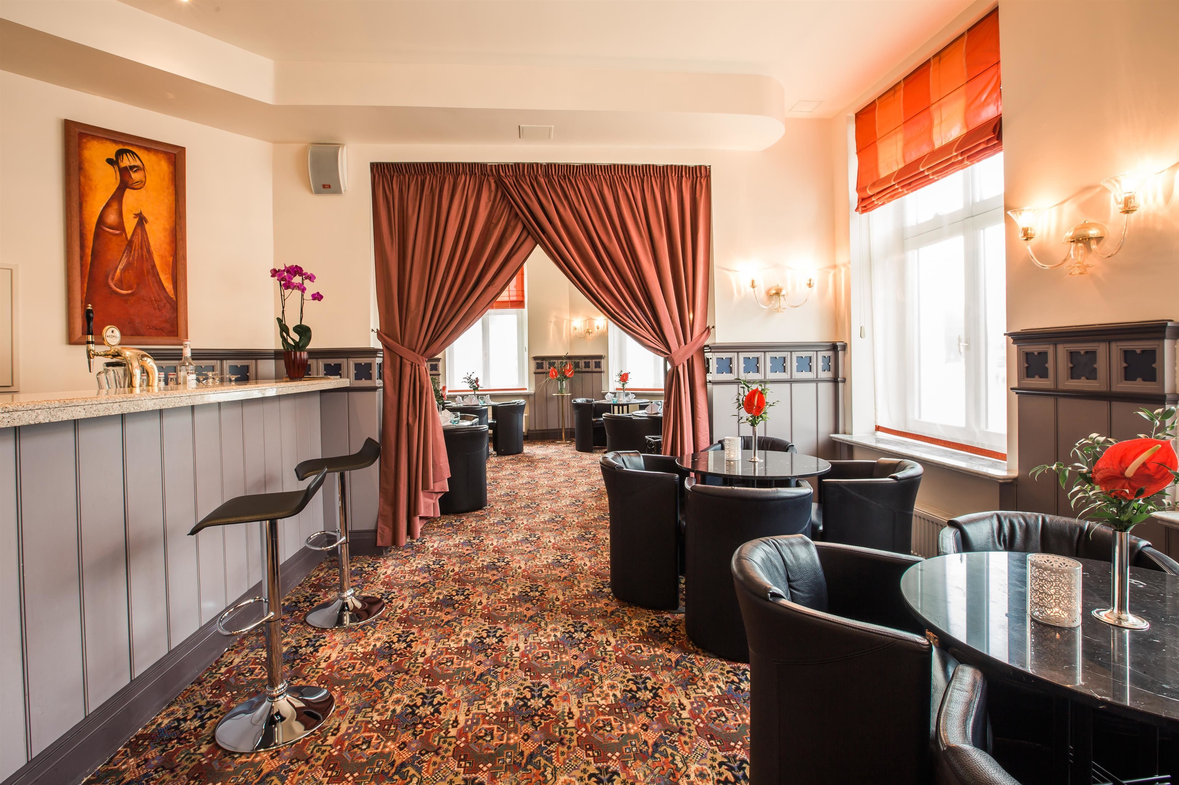 Un hall d’hôtel avec un tapis à motifs, un coin bar avec des tabourets, un espace repas avec des tables et des chaises rondes, et une grande fenêtre avec des rideaux.