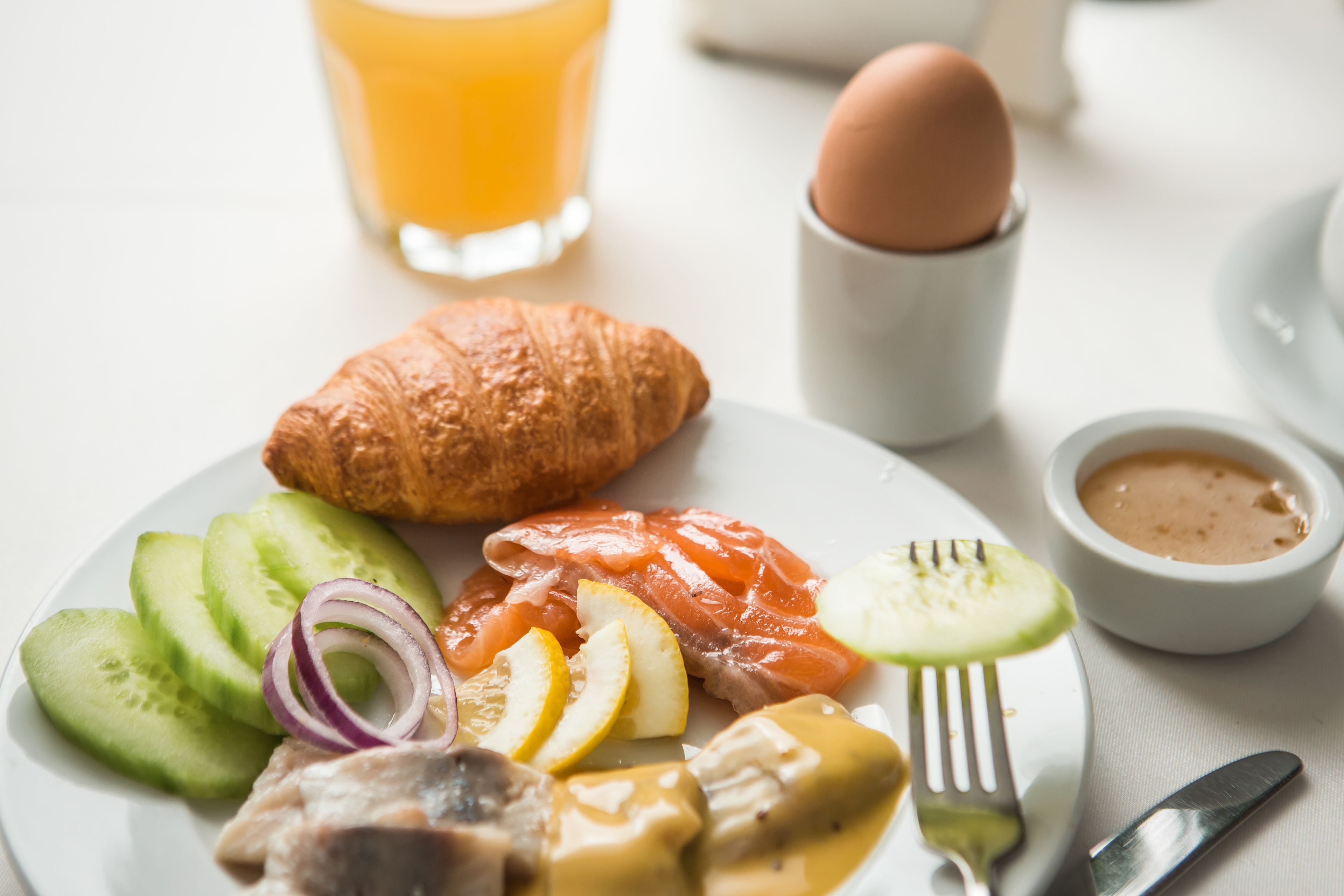 A breakfast plate with croissant, smoked salmon, cucumber slices, red onion, lemon, and a small bowl of sauce.
