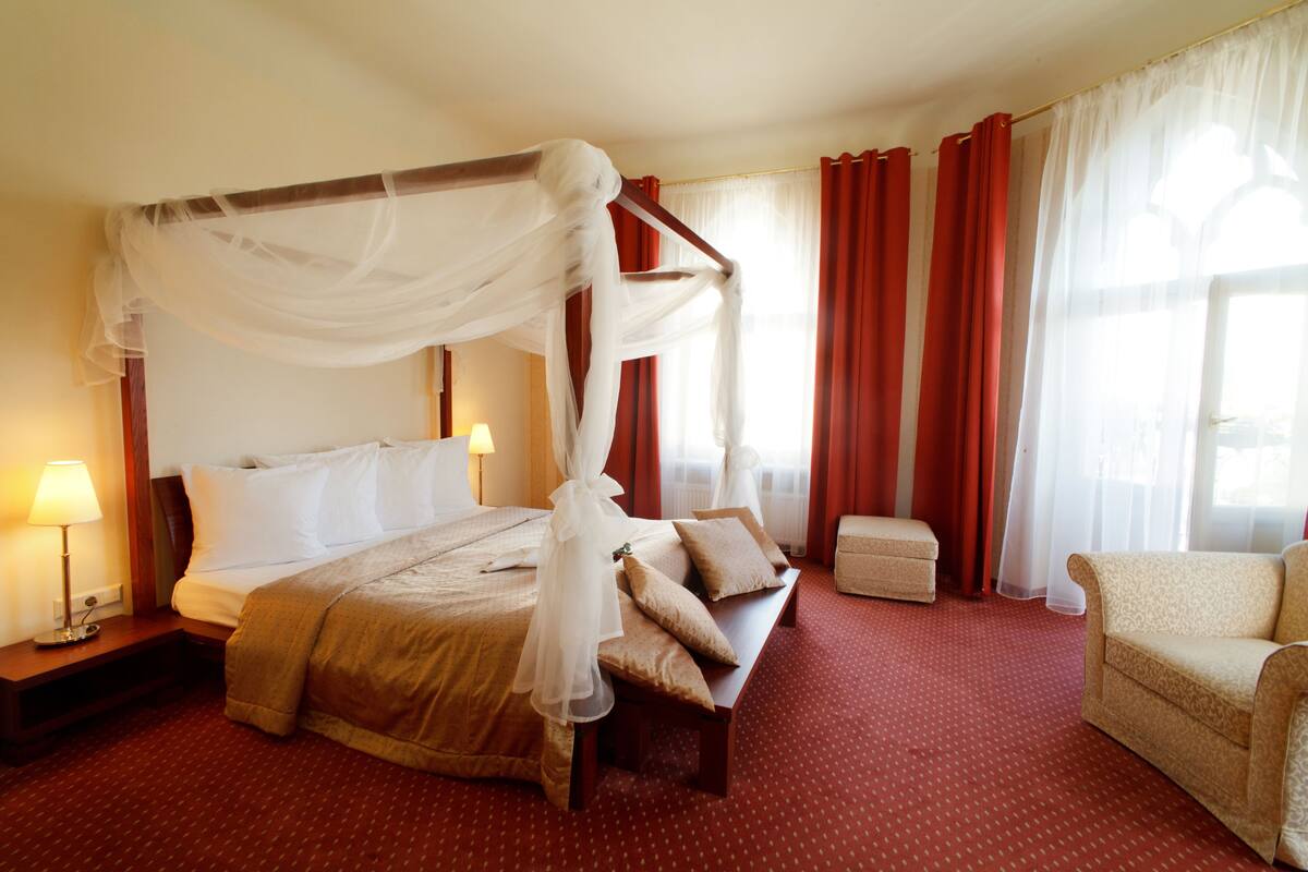 Ein Hotelzimmer mit Himmelbett, Nachttisch mit Lampe, Sofa und kleinem Hocker, mit roten Teppichböden.