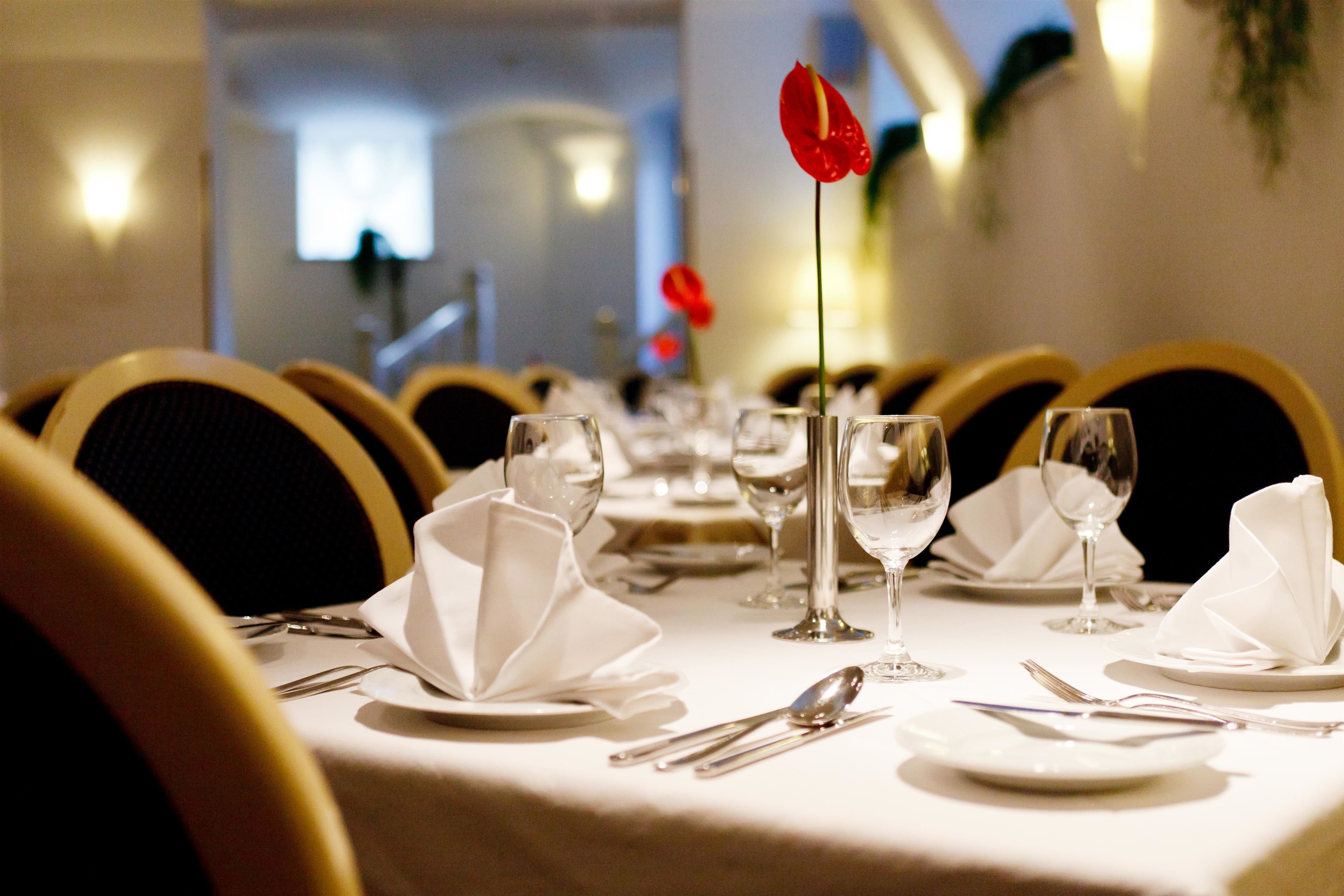 A dining table set with white tablecloths, silverware, and wine glasses, with a red flower on a napkin.