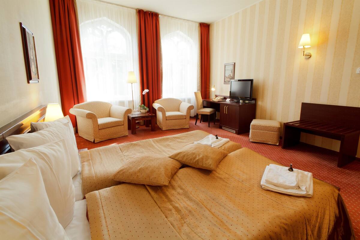 Ein Hotelzimmer mit einem großen Bett, zwei Sesseln, einem Schreibtisch inklusive Computer, einem Fernseher und einem kleinen Tisch mit Lampe.