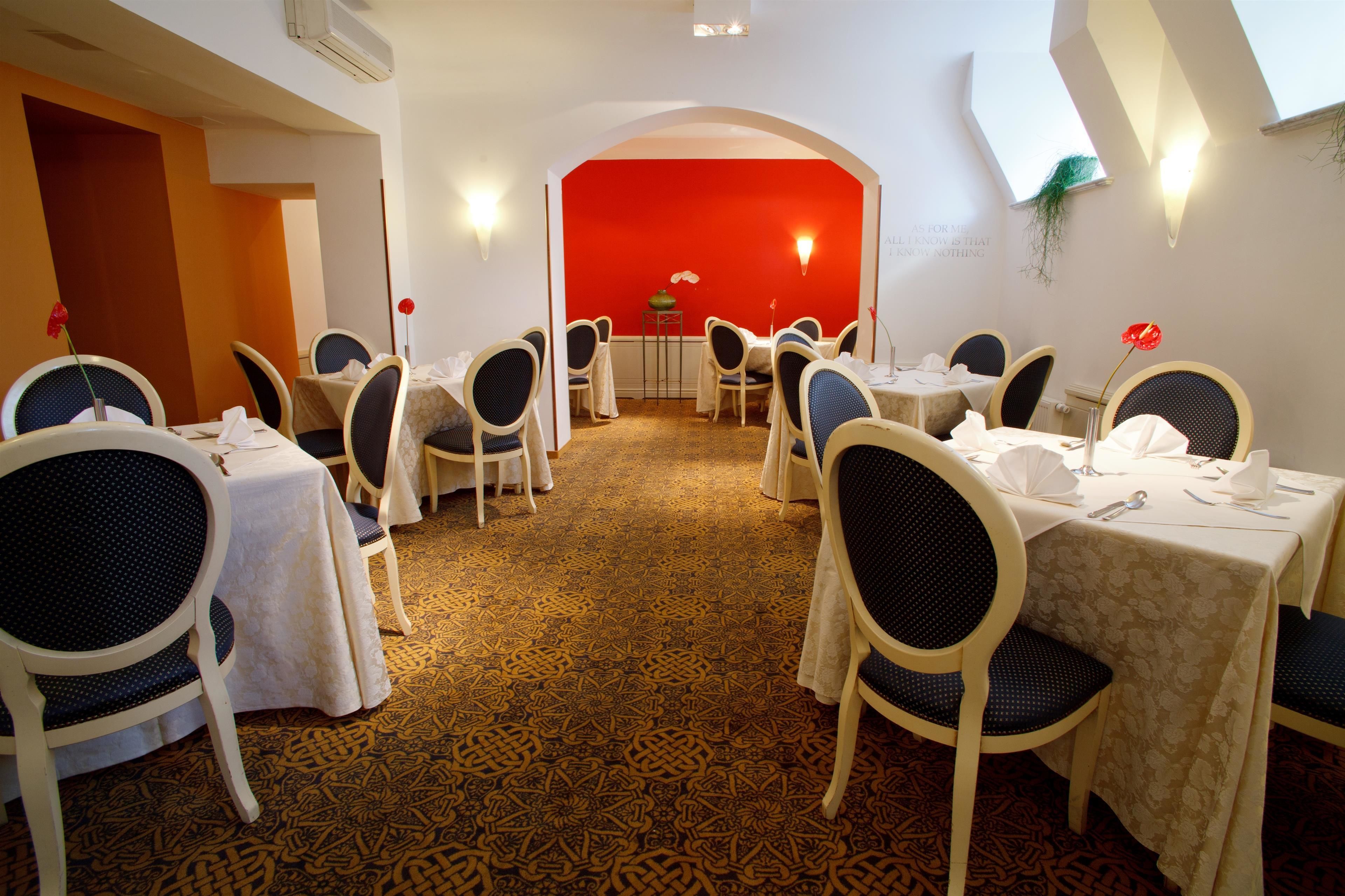A dining area with tables set for six, each with chairs and tablecloths. The room has a patterned carpet and a red accent wall.
