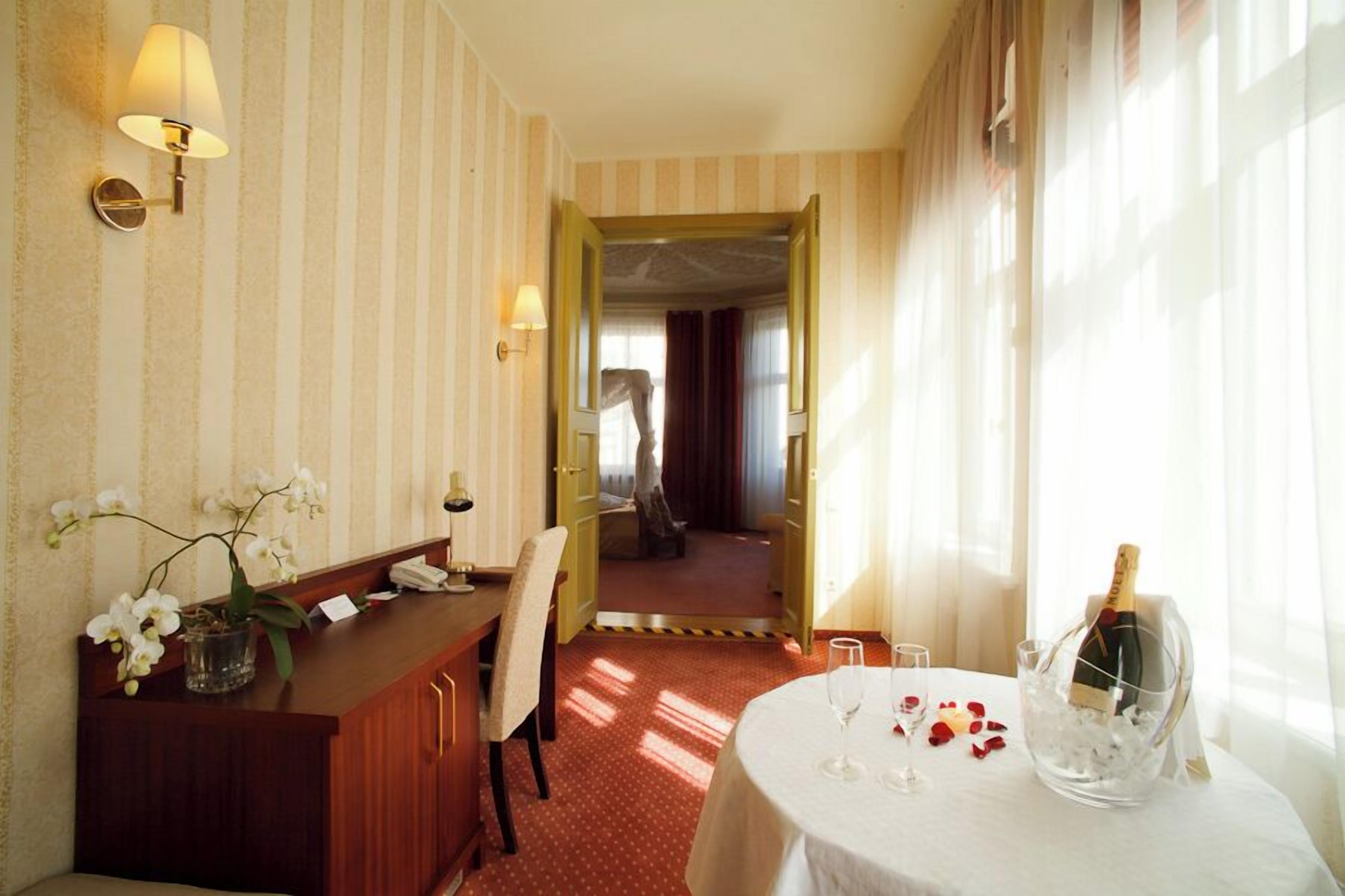 Habitación de hotel con un mostrador de recepción, un área de comedor con una mesa para dos, y un gran ventanal con cortinas.
