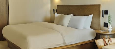 Una cama bien hecha con cabecera de madera, dos mesitas de noche con lámparas y una mesita auxiliar con una planta y un bolígrafo.