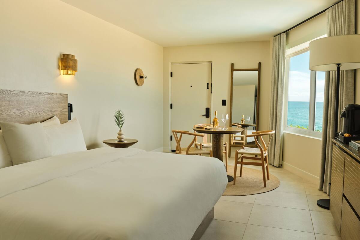 A hotel room with a bed, a dining table with chairs, a small side table with a plant, a mirror, and a view of the ocean.