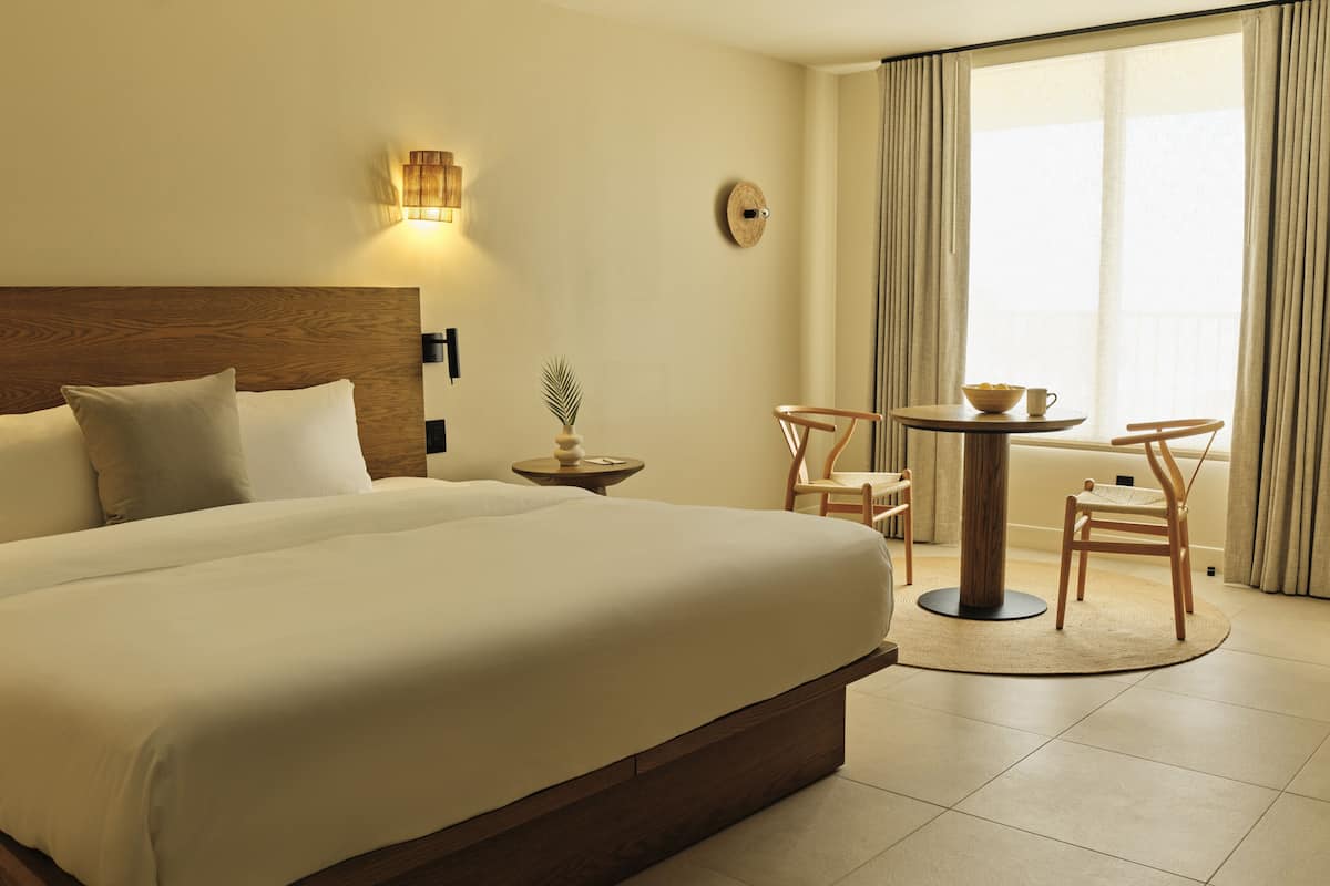 A hotel room with a large bed, a round table with chairs, a small side table, and a wall-mounted lamp.