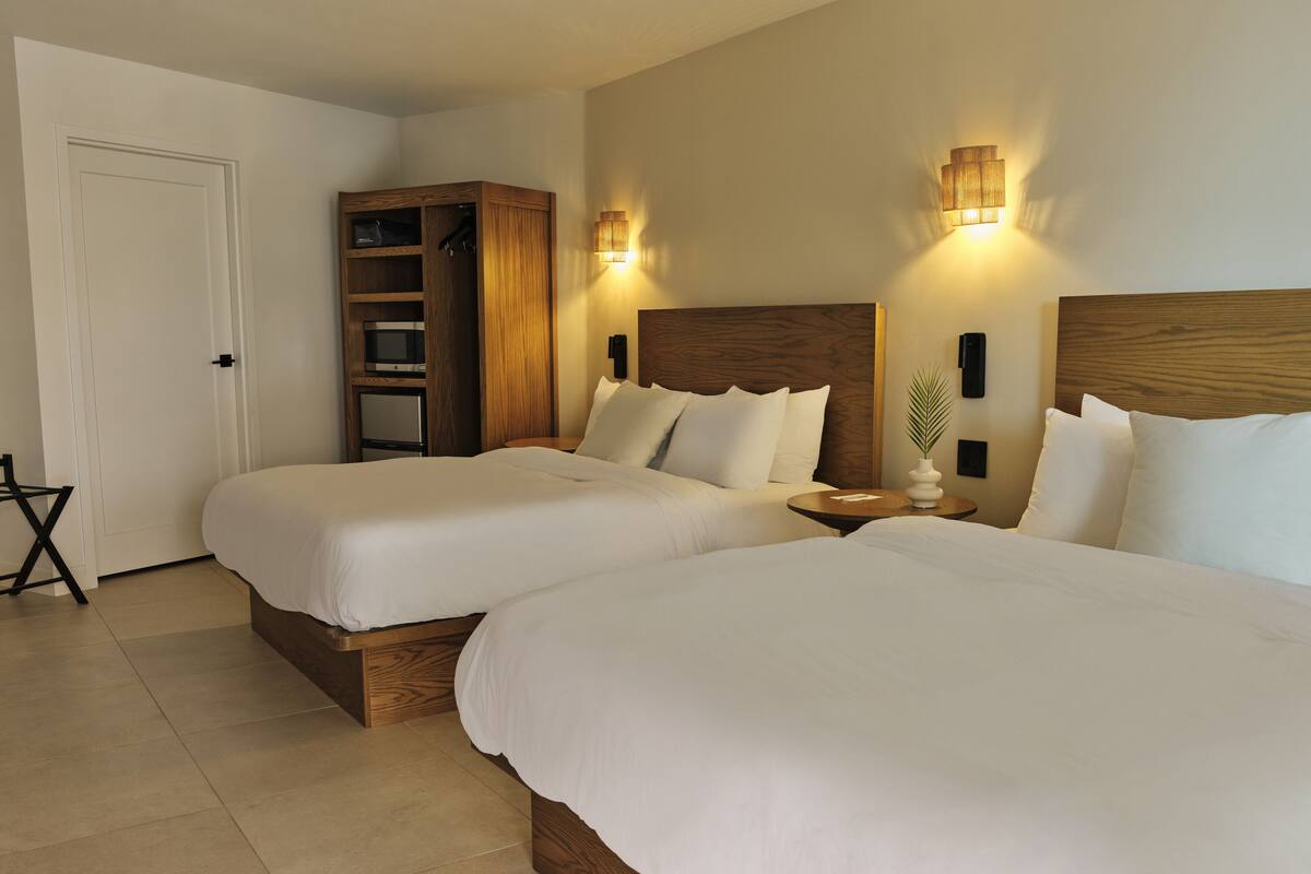 A hotel room with two beds, a wooden wardrobe, a small table, and a wall-mounted light fixture.