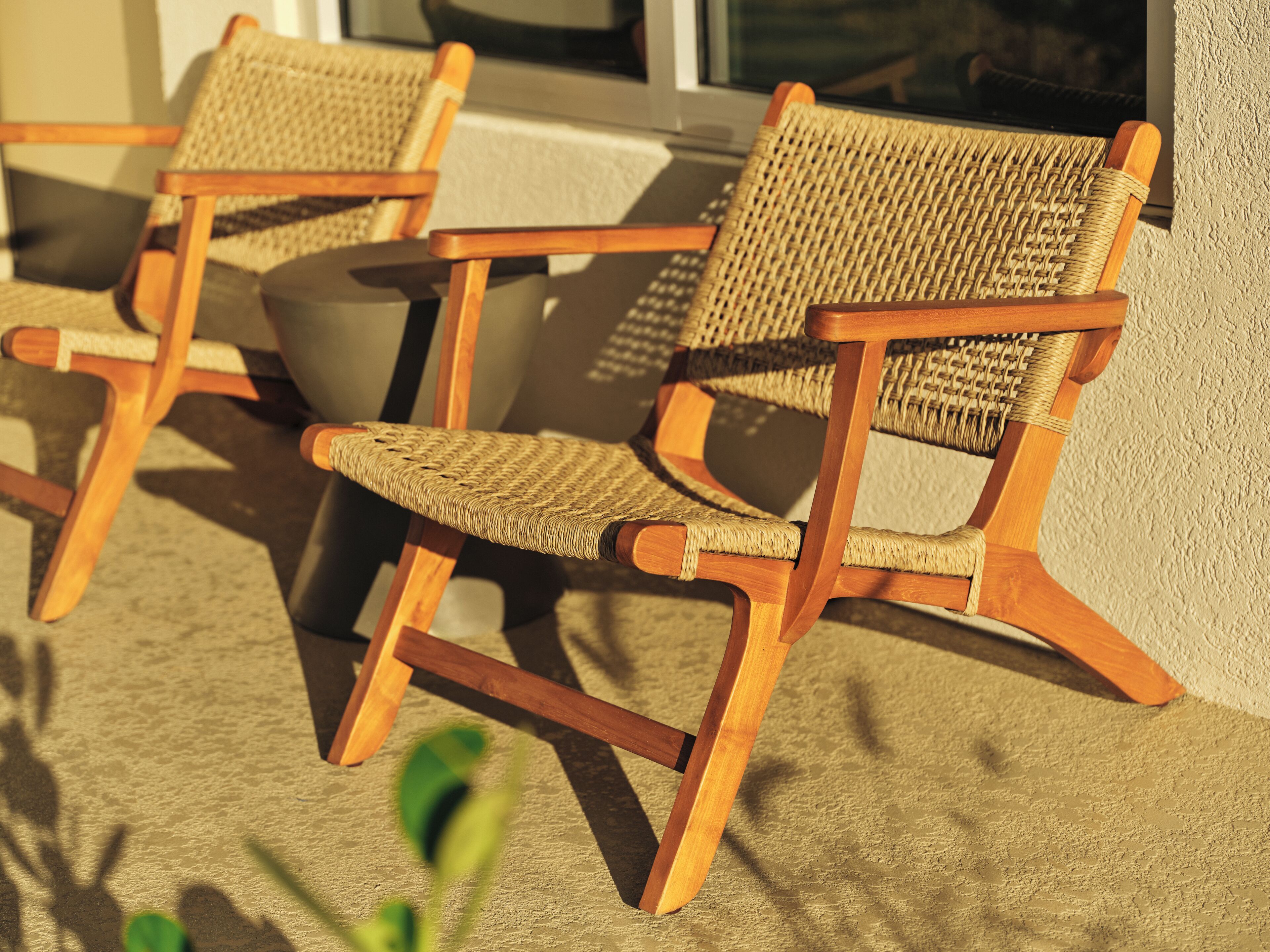 Two wicker chairs with wooden frames positioned on a textured surface, likely a patio or deck, with a building in the background.