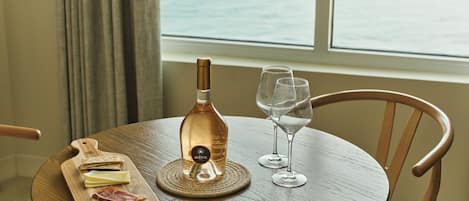 A round wooden table with a bottle of wine, two wine glasses, a cheese board with cheese and ham, and a corkscrew on a woven mat.