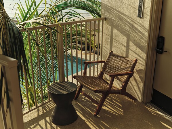A balcony with a wicker chair and a small round table, overlooking a swimming pool and palm trees.