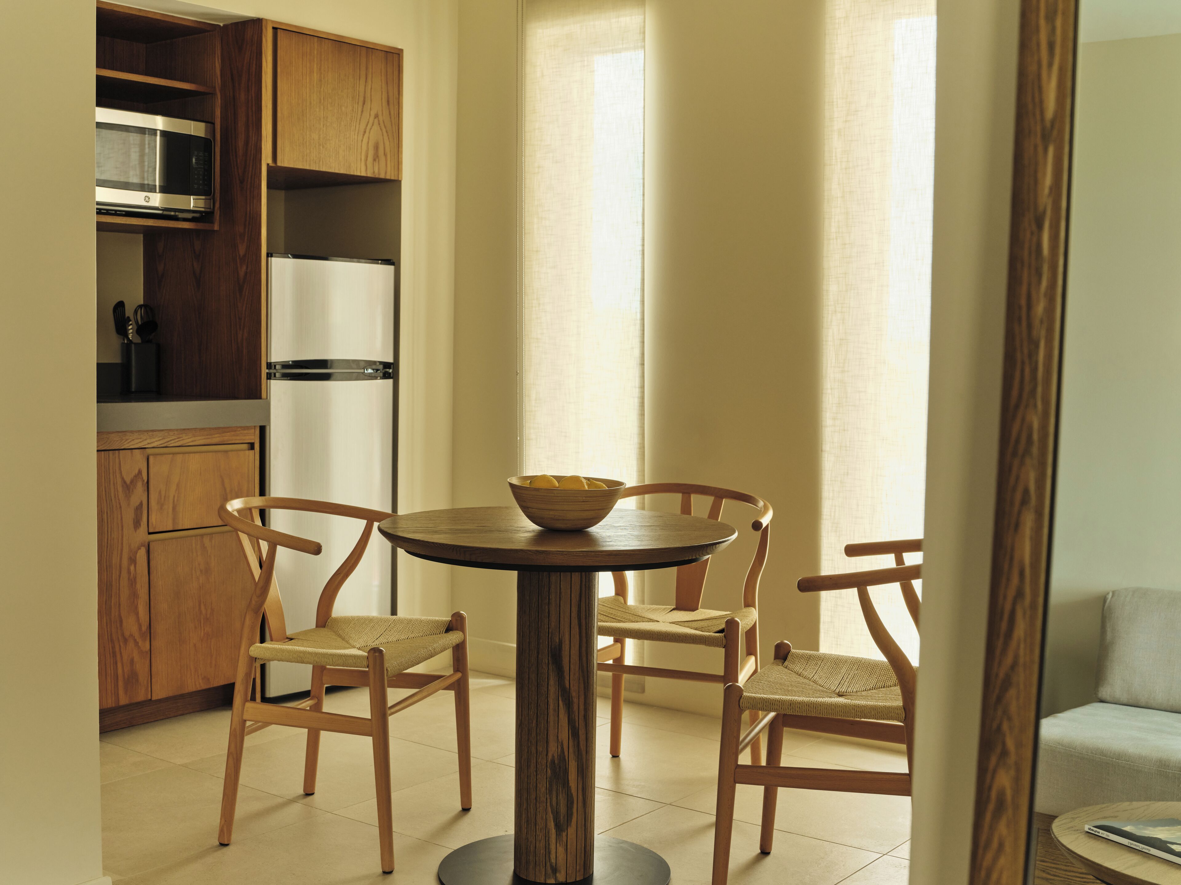 Una cocina moderna con armarios de madera, un microondas y un refrigerador. Hay una mesa redonda de madera con dos sillas y un bol de fruta.