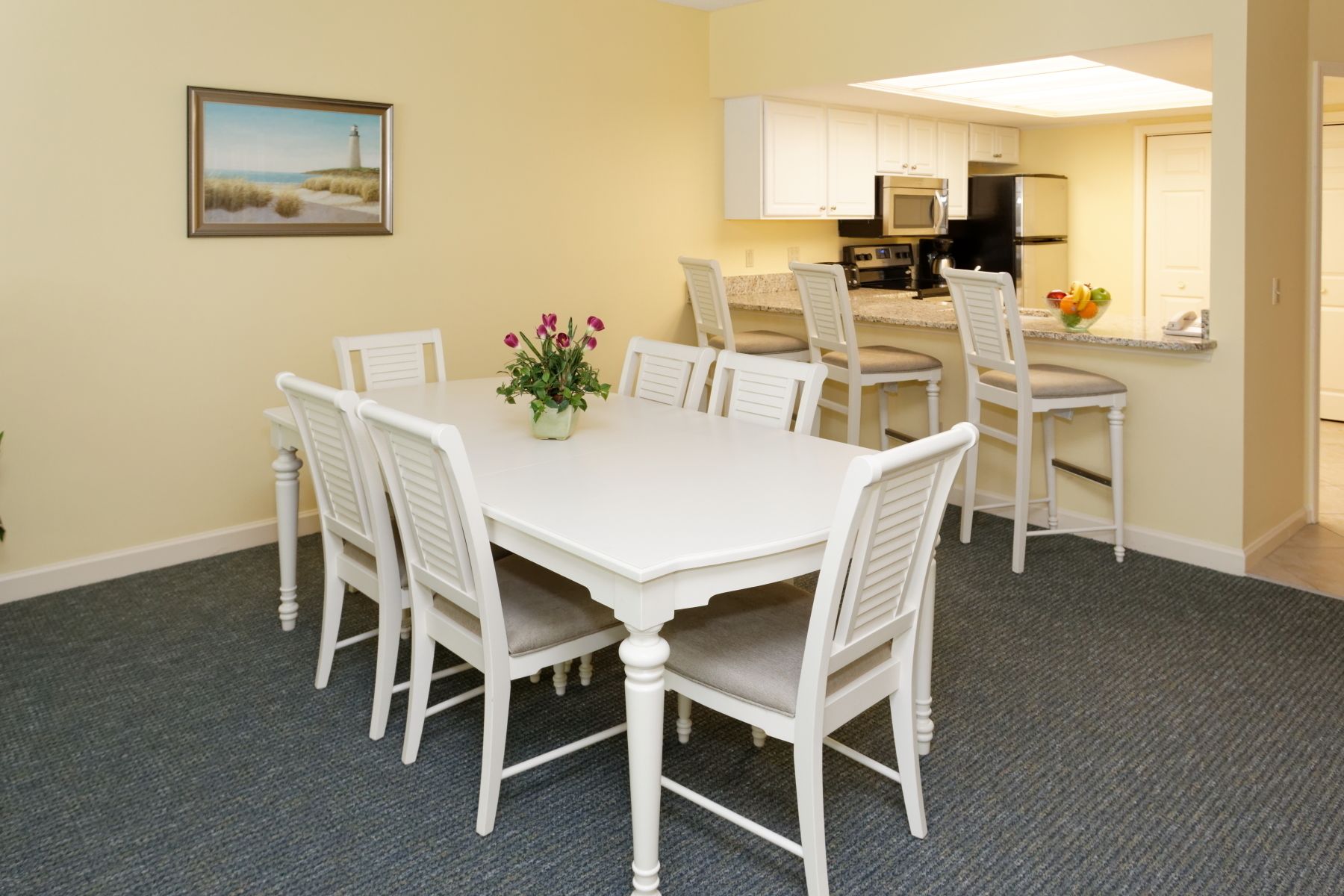 A dining area with a white table and chairs, a painting on the wall, and a kitchen in the background.