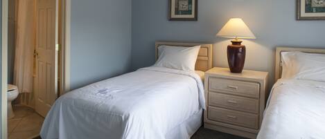 A hotel room with two beds, a nightstand with a lamp, a bathroom with a toilet and shower, and two framed pictures on the wall.